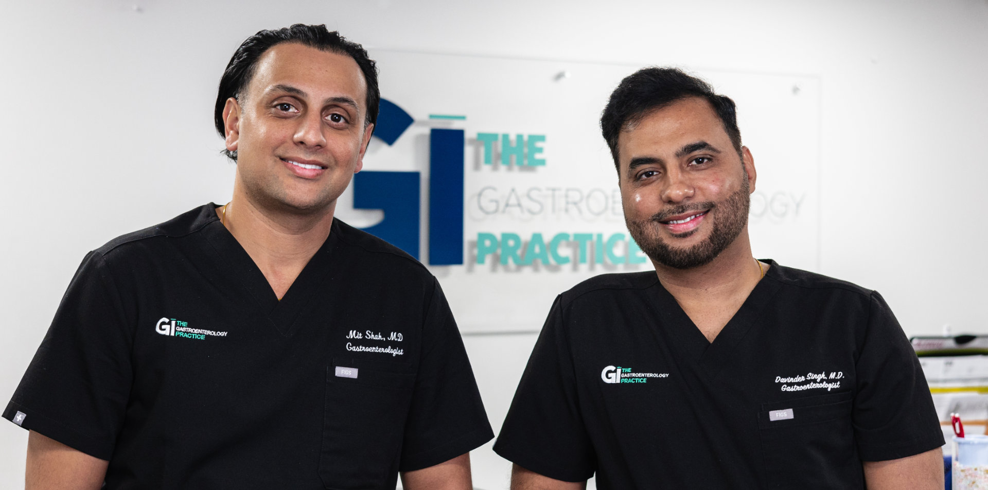 Two men in black medical scrubs stand side by side, smiling, in a clinical office with a sign reading "The Gastroenterology Practice" in the background.
