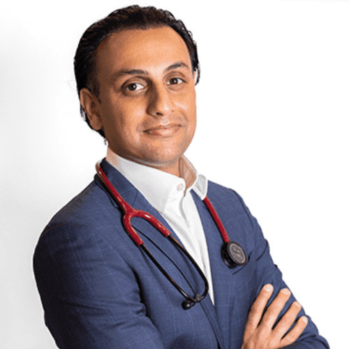 A man in a blue suit with a stethoscope around his neck stands with arms crossed against a plain white background.