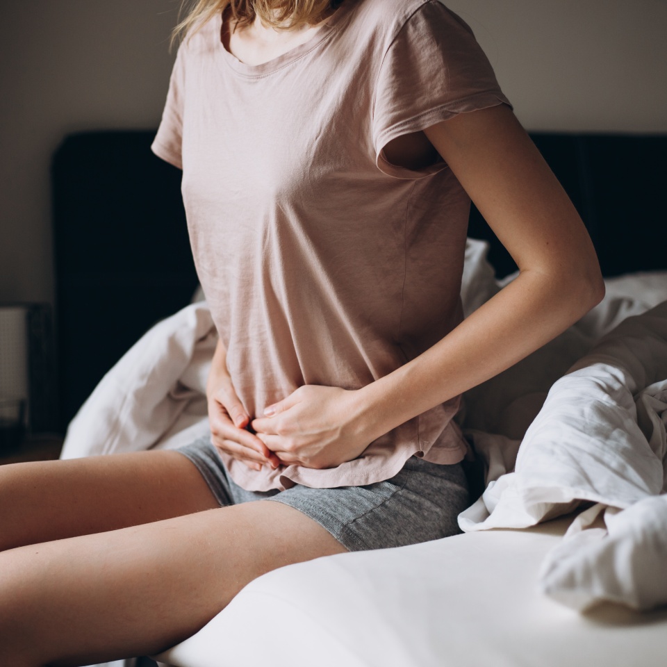 Young woman suffering from strong abdominal pain, casual style indoor shoot. Female sitting on bed in morning