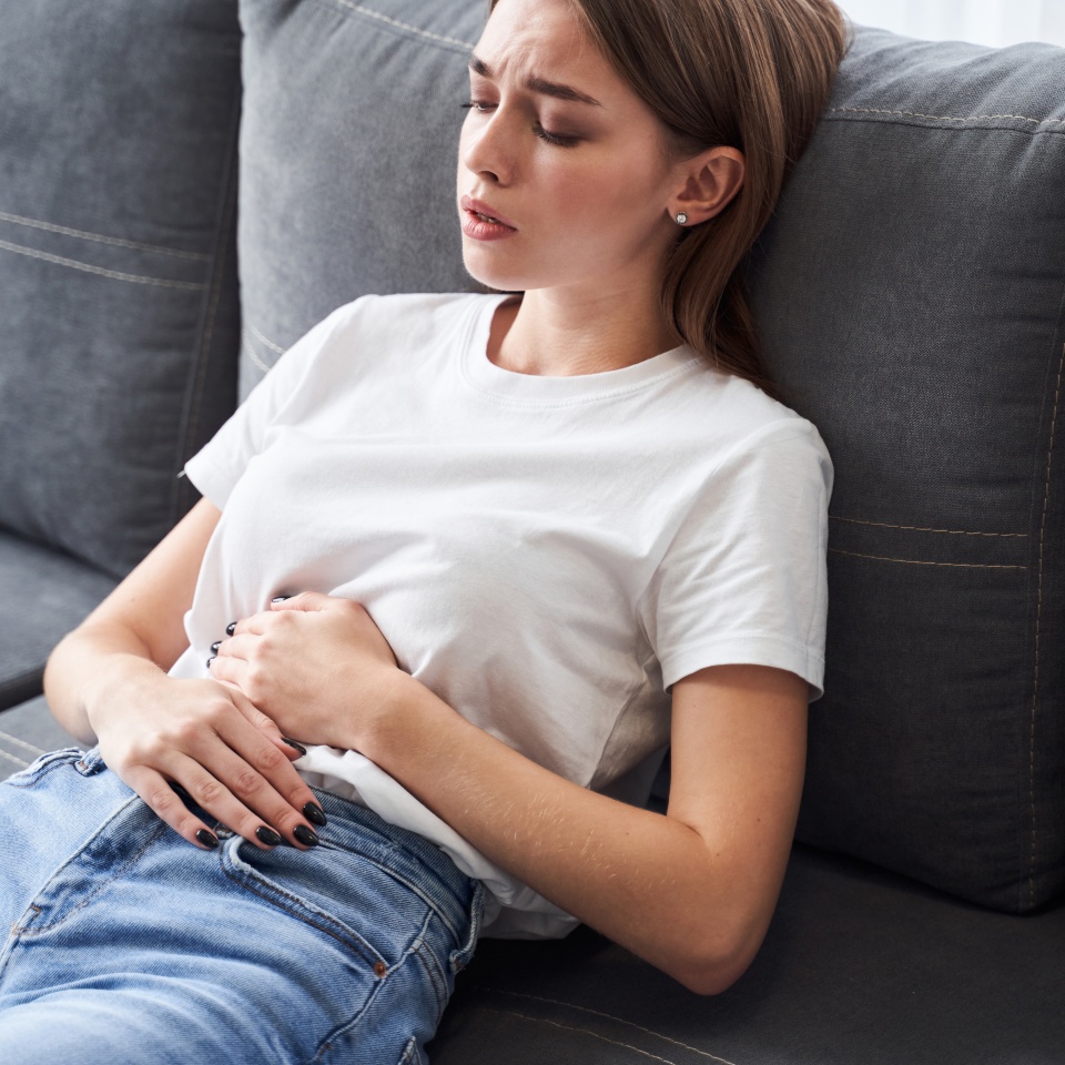 Female pain. Woman with pain in stomach is sitting on couch at home. Young woman suffering from strong abdominal pain while sitting on sofa at home