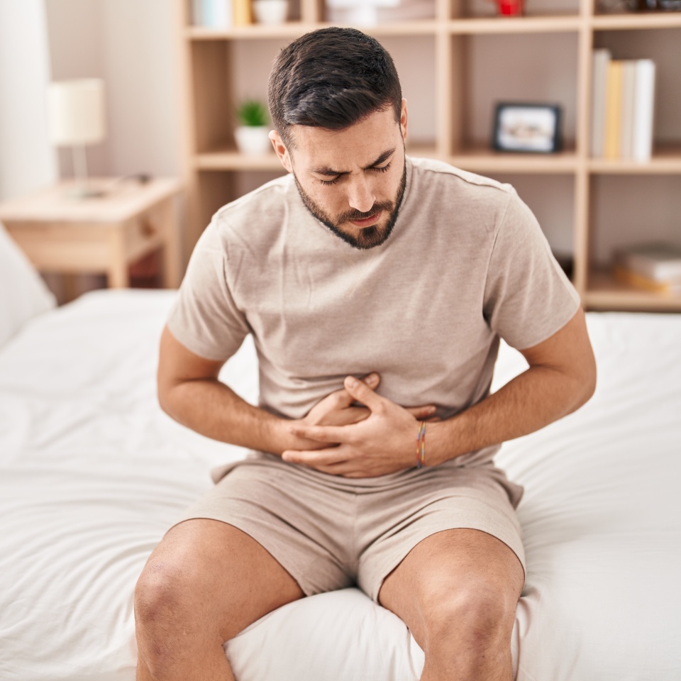 Young hispanic man suffering for stomachache sitting on bed at bedroom