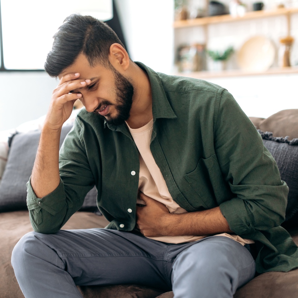 Unhappy indian or arabian man, sits on comfortable sofa in cozy living room, holds his hands on his stomach, grimaces from pain in his stomach, suffers from poisoning, spasm, stomach problems
