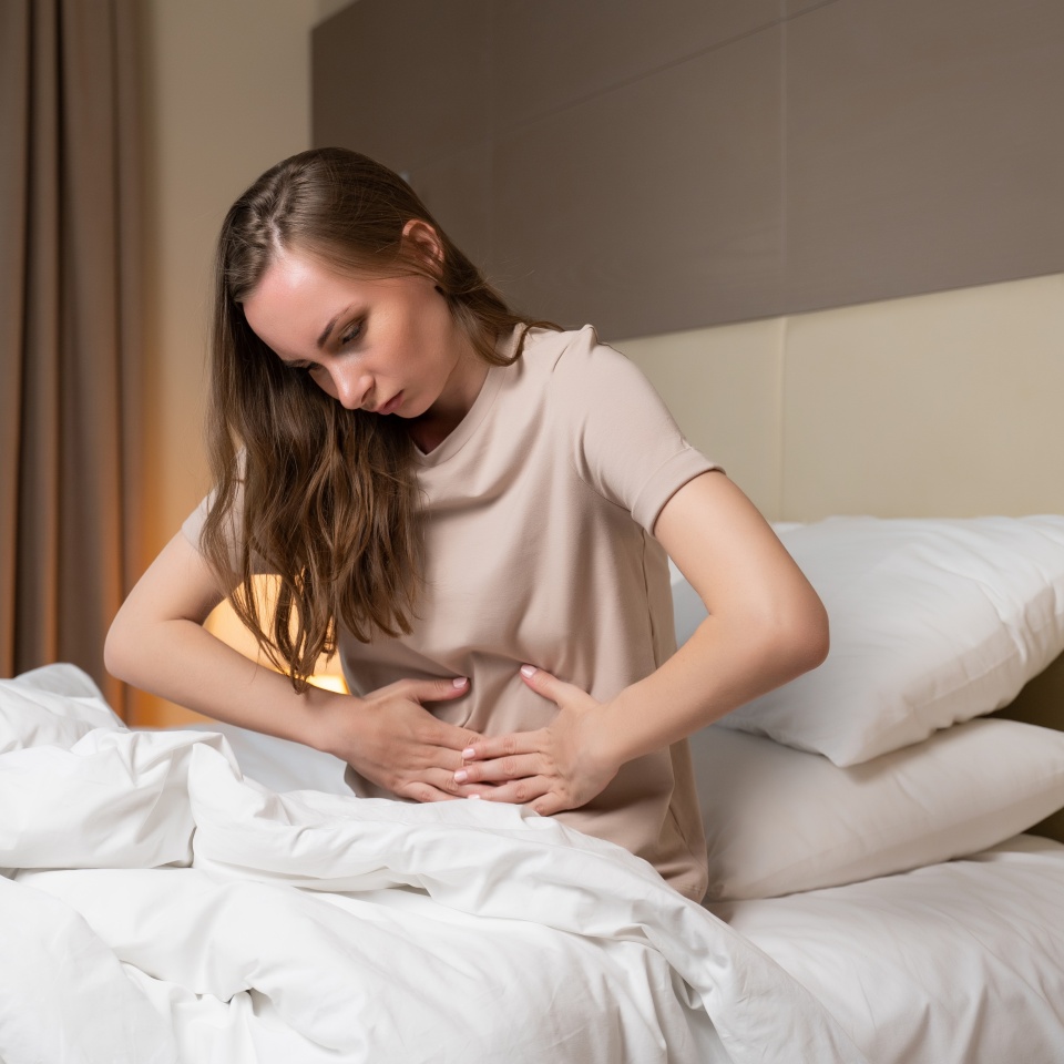Woman curls up touching stomach with hands sitting on bed in hotel room. Sick female person feels pain in abdomen after waking up in early morning