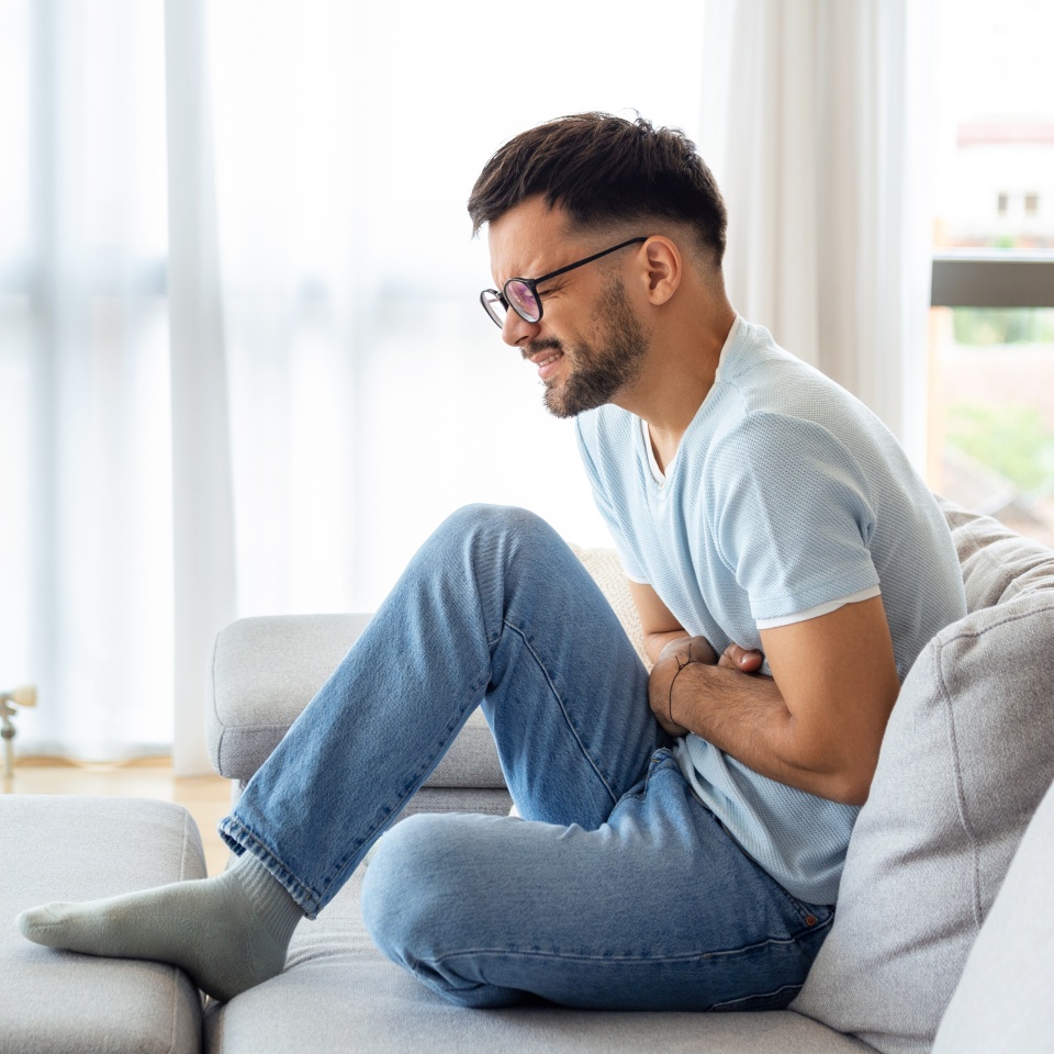 A man suffers from abdominal pain while sitting at home on the couch. Young man suffering from abdominal pain sitting on the couch at home.