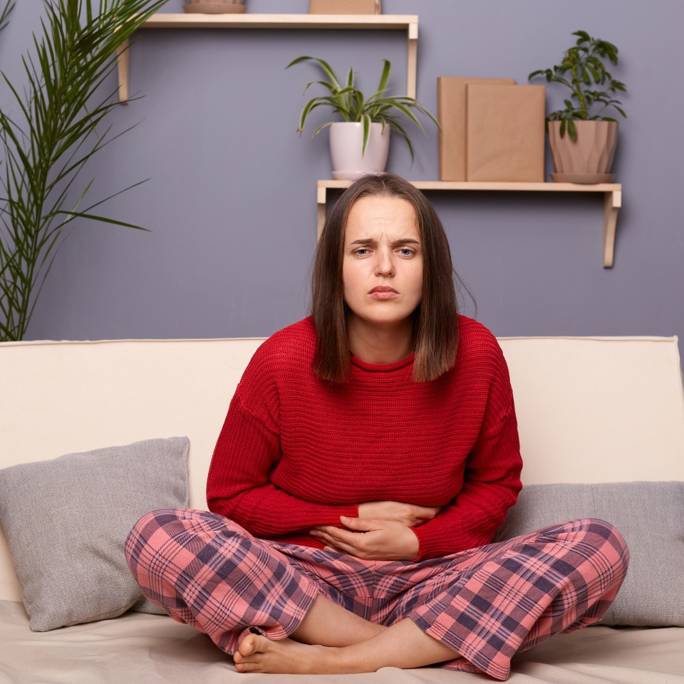 Horizontal shot of sick woman wearing red sweater and checkered pants sitting with crossed legs on sofa in home interior, touching her belly, suffering period pain or has food poisoning.