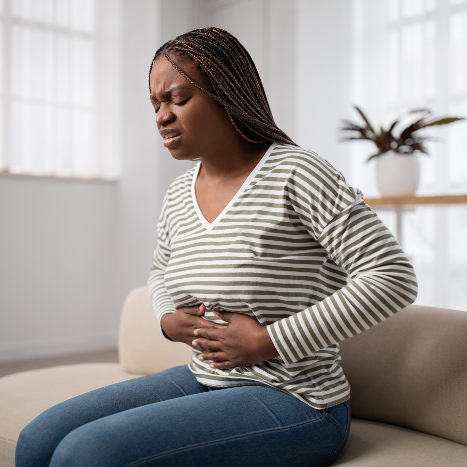 Sad young african american woman wearing casual clothes suffering from menstrual pain, feeling sick to her stomach, holding belly, having abdominal cramps during period, sitting on couch at home