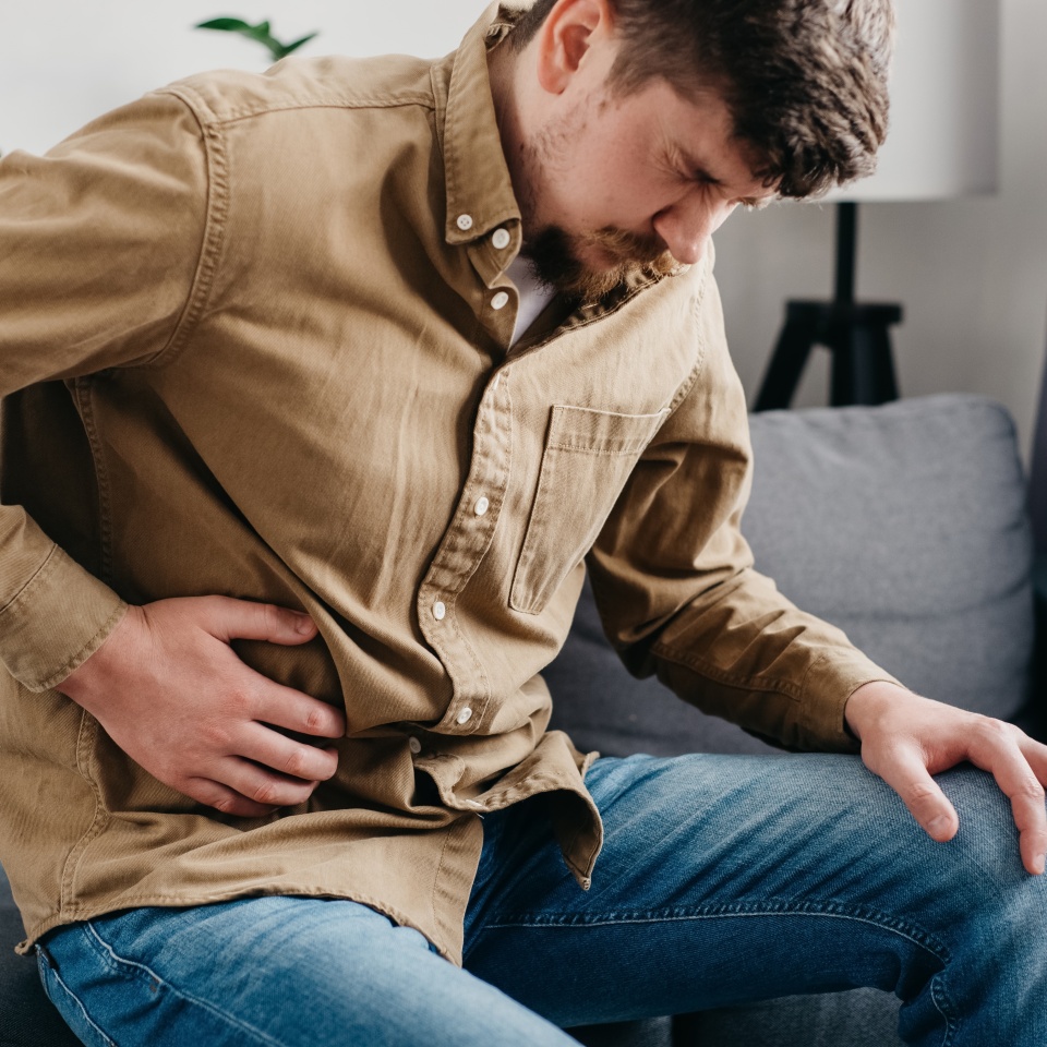 Unhealthy young caucasian man 30s holding belly sit on couch at home feel bad digestive problems abdominal pain illness suffers from liver damage bending body in ache feeling unwell poisoning concept