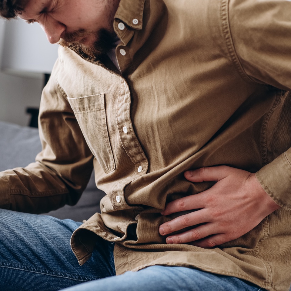 Upset male squeezing belly with hand because of abdominal pain. Unhappy young man sit on grey couch at home, suddenly feeling strong stomach ache, gastritis problem. Guy suffering from stomach ache