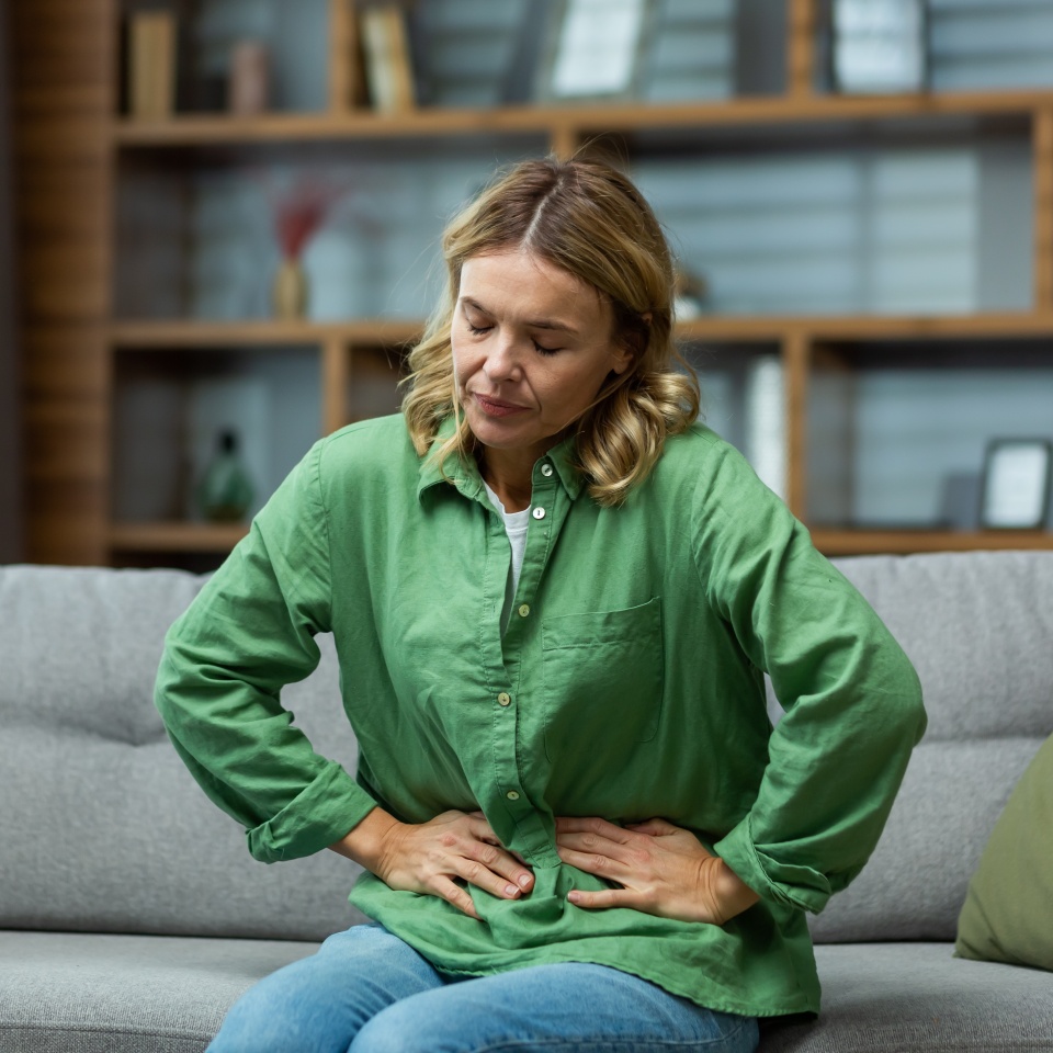 An elderly woman is sitting on the sofa at home, holding her stomach with her hands. Feels the pain of internal organs, indigestion, poisoning, menstrual pain.