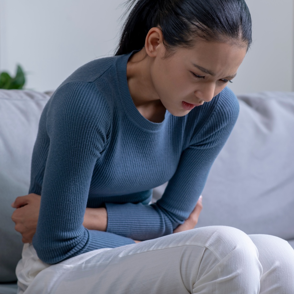 Young Asian woman suffering from strong abdominal pain while sitting on sofa at home. Sick female having a stomach ache.