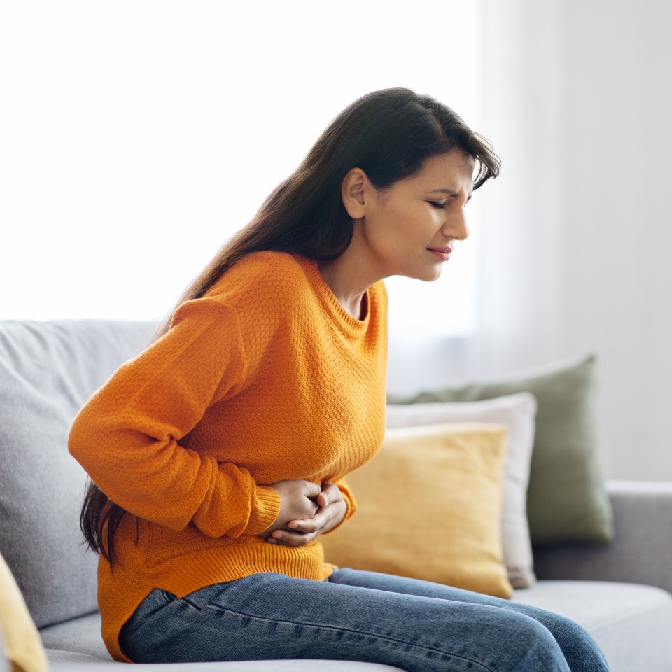 Unhappy sick pretty long-haired young indian woman wearing casual comfy outfit sitting on couch in living-room at home, touching belly, suffering from period crumps, side view, copy space
