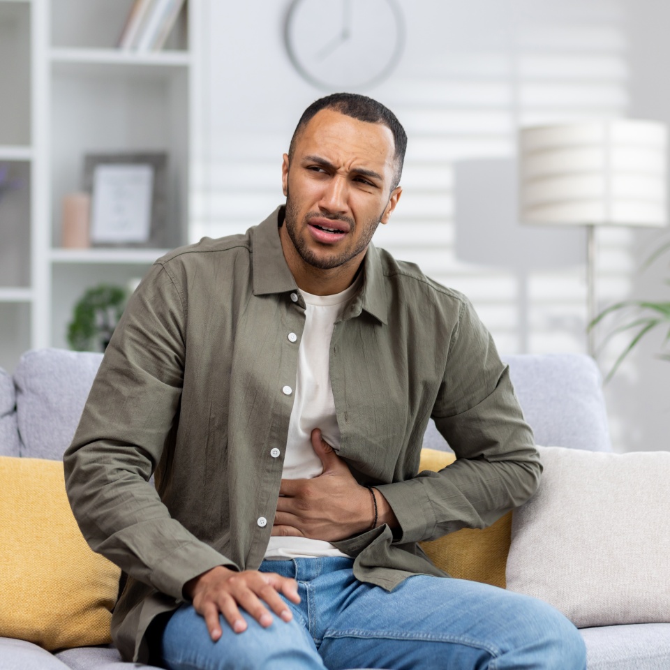 A young African American man is suffering from abdominal pain, stomach cramps and constipation. He is sitting on the sofa and holding his stomach, grimacing in pain.
