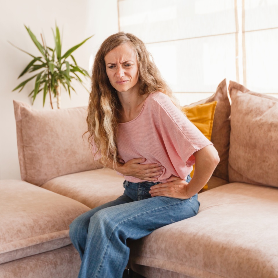 Young woman suffering from strong abdominal pain while sitting on sofa at home. Woman having painful stomach ache