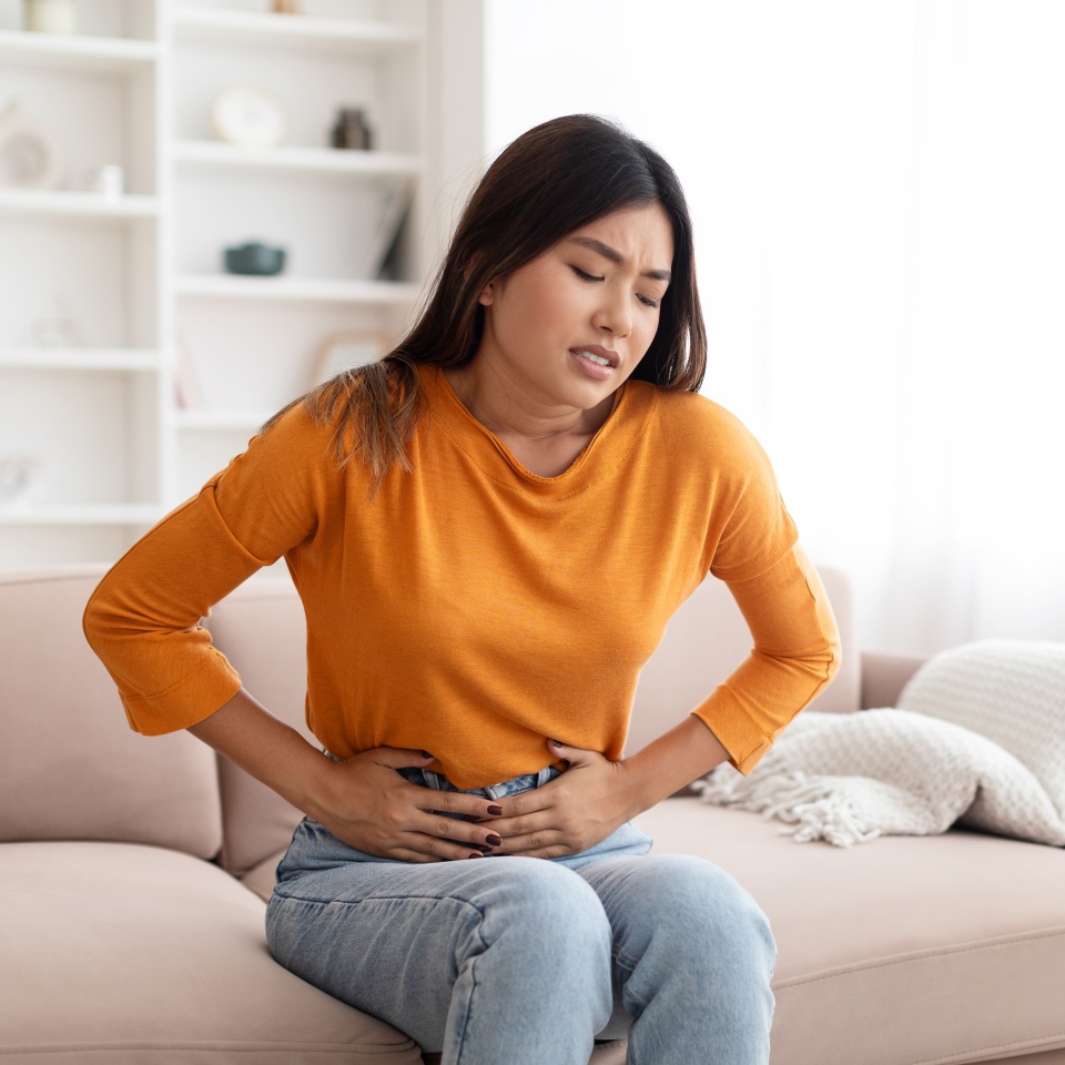 Unhappy sick pretty long-haired young asian woman wearing casual comfy outfit sitting on couch in living-room at home, touching belly, suffering from period cramps, copy space