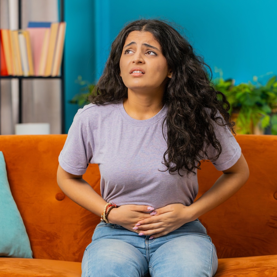 Girl holding belly, feeling abdominal menstrual pain. Sick ill Indian woman suffering from period cramps, painful stomach ache on sofa at home room. Abdominal pain, gastritis, diarrhea, indigestion