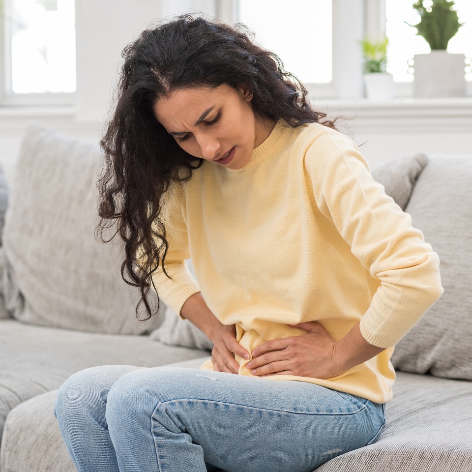 Portrait of a young woman sitting on a sofa in the living room suffering from sharp pain in the lower abdomen. The girl holds her stomach with her hands and suffers from menstrual pain or nausea.
