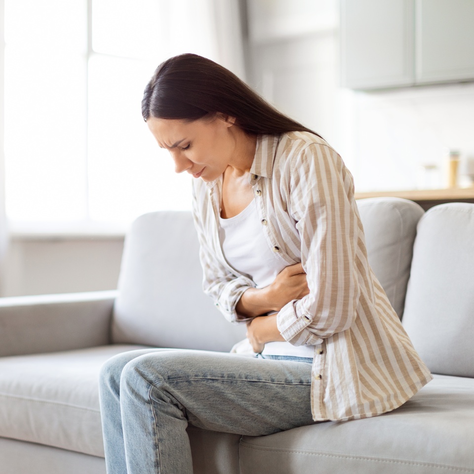 Digestion Problems. Portrait Of Sick Young Woman Suffering From Acute Abdominal Pain At Home, Upset European Female Having Menstrual Pain Or Stomach Ache, Sitting On Couch And Touching Belly
