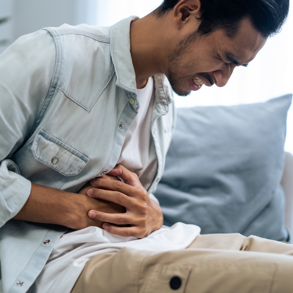 Asian attractive sick male suffer from hurt stomachache in living room. Handsome young man sitting on sofa, feeling bad and painful having stomach ache then clasping belly in house. Health Problem.