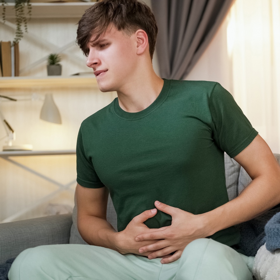 Stomach pain. Indigestion suffering. Unhappy man with abdomen problem feeling sick grasping belly in cramps sitting on sofa in home living room interior.