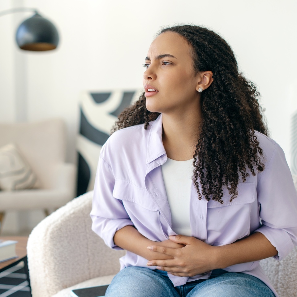 Unhappy sick young curly haired brazilian or hispanic woman in casual clothes, sitting on couch in a living room at home, touching belly, suffering from period cramps, stomachache, feels pain
