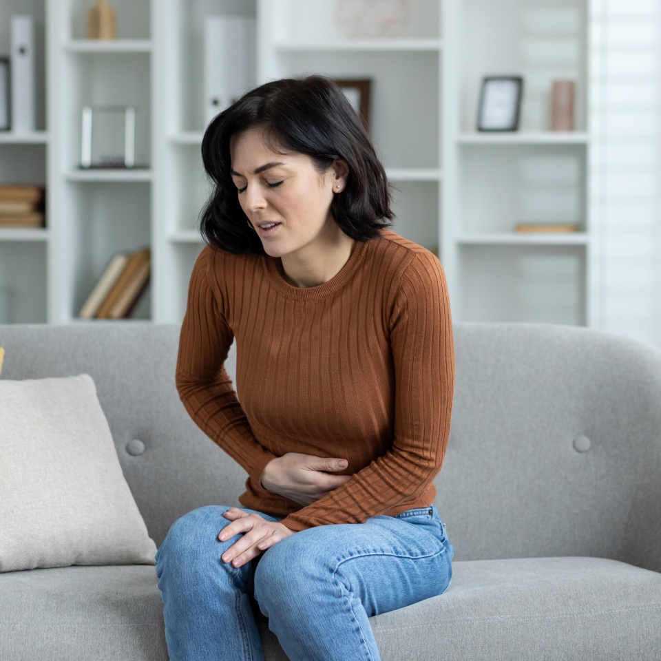 A young woman grimacing in pain is sitting on the sofa at home and holding her stomach with her hand, feeling severe pain.