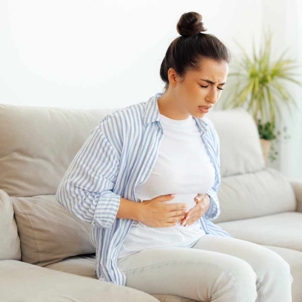 Sad arabian or indian woman sitting on a sofa, suffering stomach ache, constipation, cramp, nausea or bloated, holds her stomach with her hands, stomach pain, problems with indigestion, menstruation