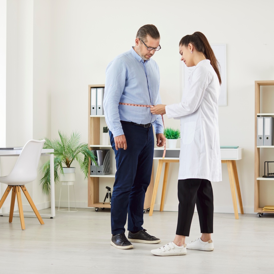 Young female doctor physiotherapist or nurse measure waist with measuring tape for mature man patient standing in medical clinic during examination. Slimming, weight loss and diet concept.