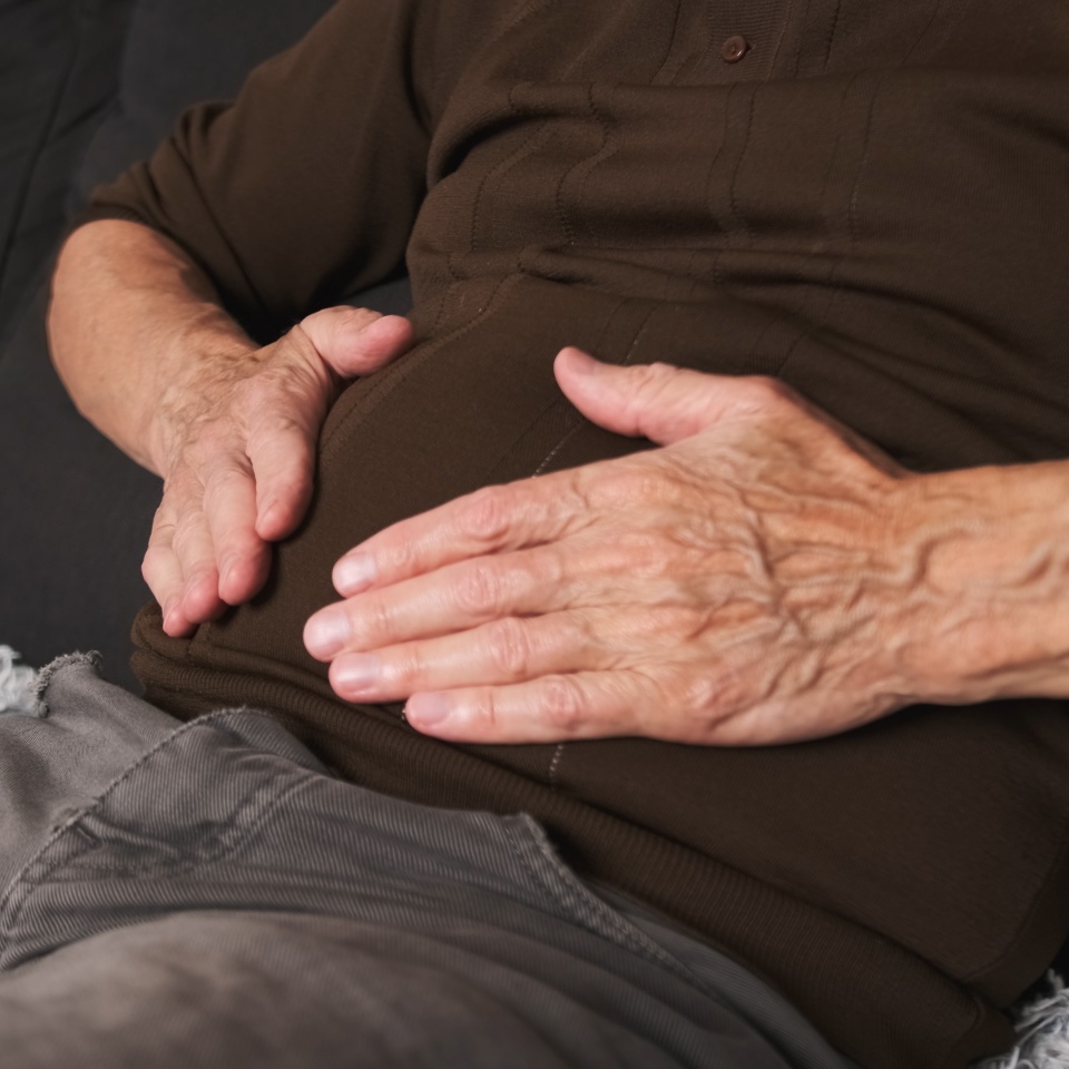 Elderly man having painful stomach ache sitting on sofa, close-up, Caucasian senior man suffering from abdominal pain, Period cramps, Hands squeezing belly