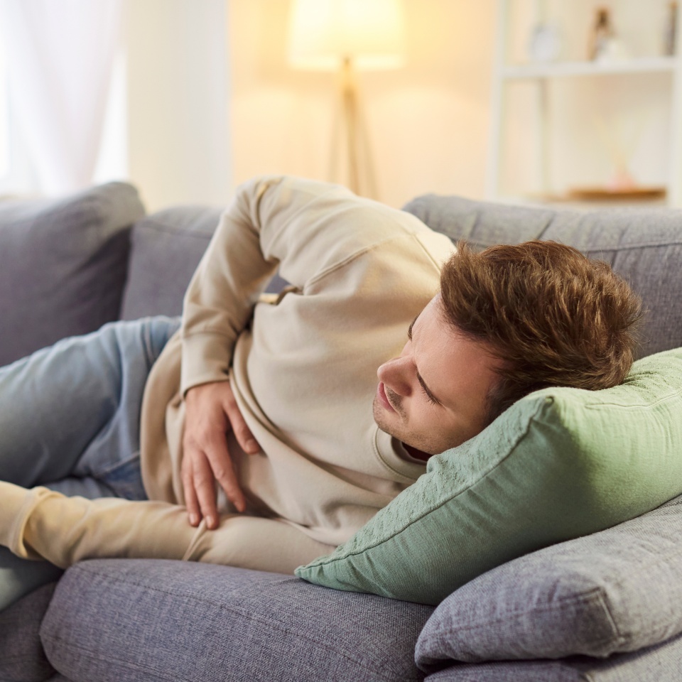 Young sick man touching his belly suffering from stomachache. Male person in casual clothes with abdominal pain lying on sofa in the living room at home. Health problem and medicine concept.