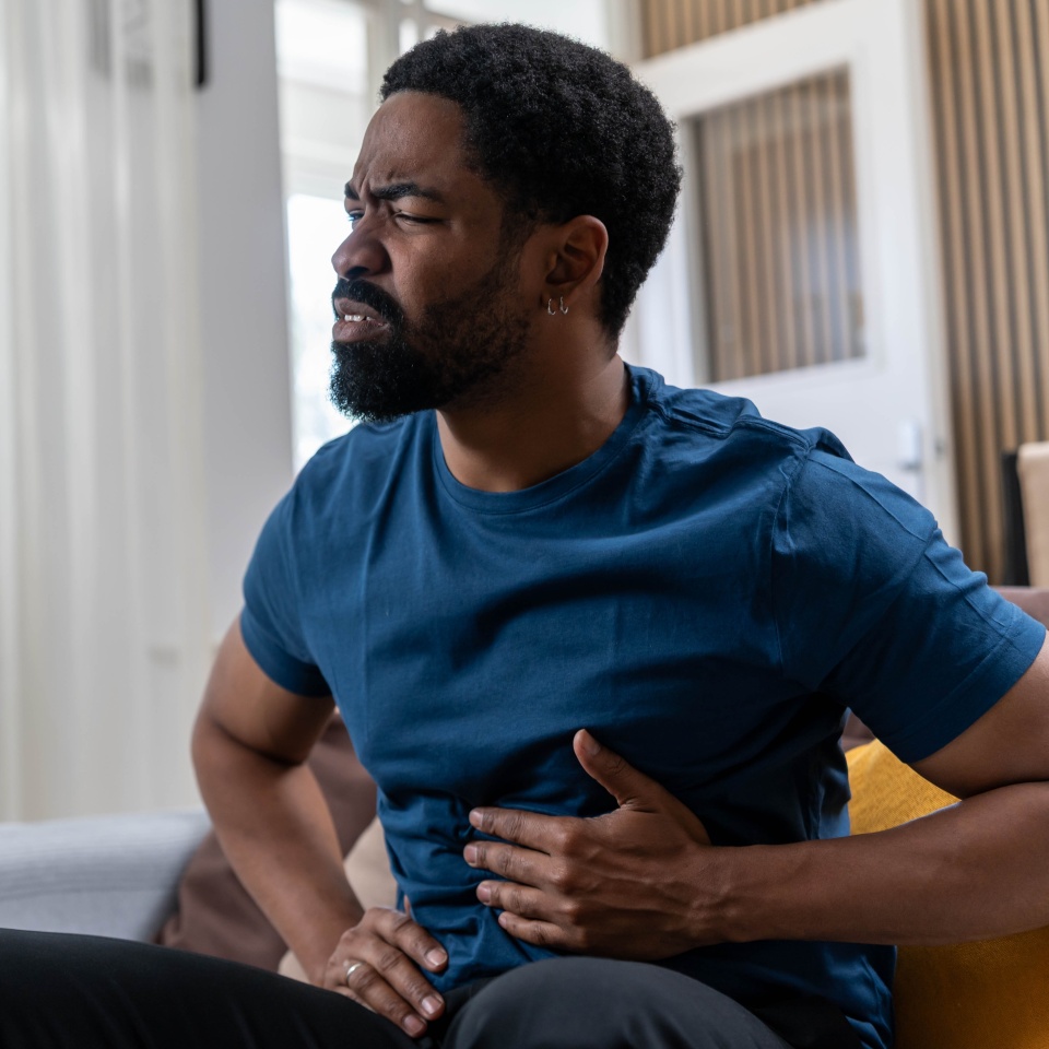 Sad young Black man suffering from strong stomach ache while sitting on couch at home during daytime. Abdominal pain, gastritis or digestive issue.