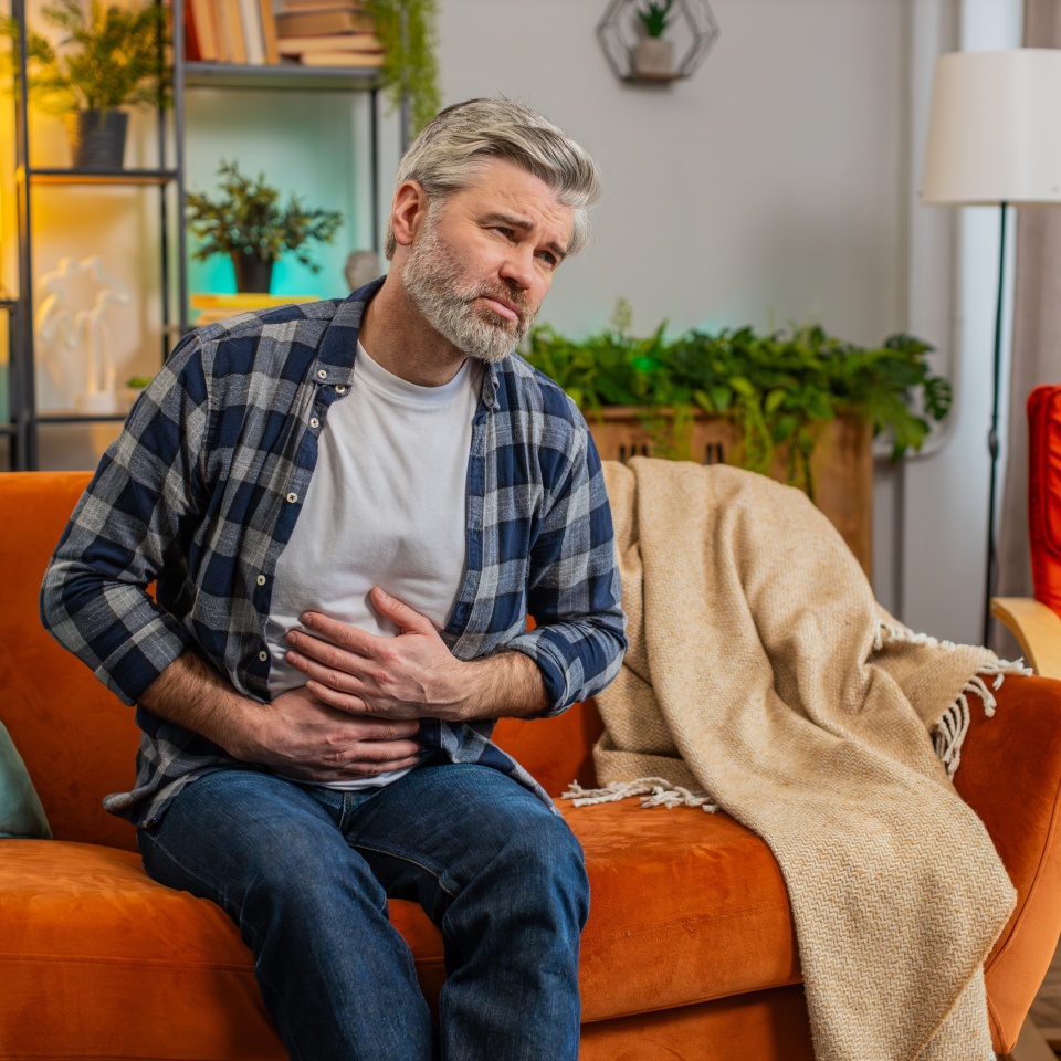 Senior bearded man sitting on couch feeling sudden strong abdominal stomach ache, gastritis problem. Middle-aged guy having symptom poisoning diarrhea indigestion peptic ulcer pancreatitis at home.