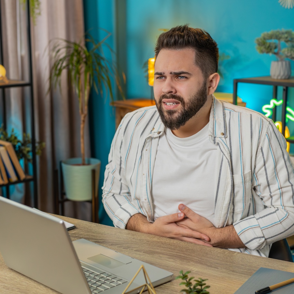 Disappointed freelancer man feeling overeating during work process on laptop in home office. Upset businessman freelancer guy man touching stomach and experiencing discomfort and sharp pain. Lifestyle