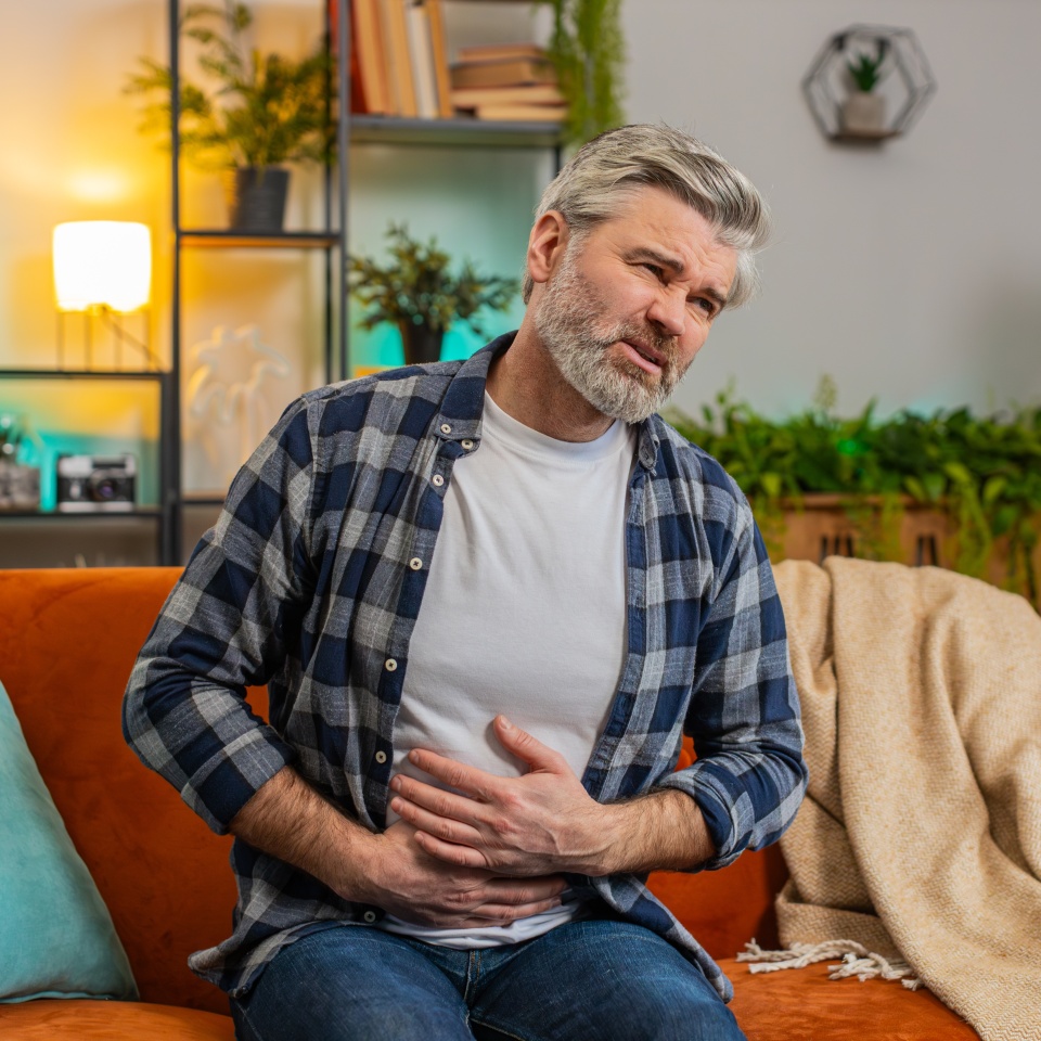 Senior bearded man sitting on couch feeling sudden strong abdominal stomach ache, gastritis problem. Middle-aged guy having symptom poisoning diarrhea indigestion peptic ulcer pancreatitis at home.