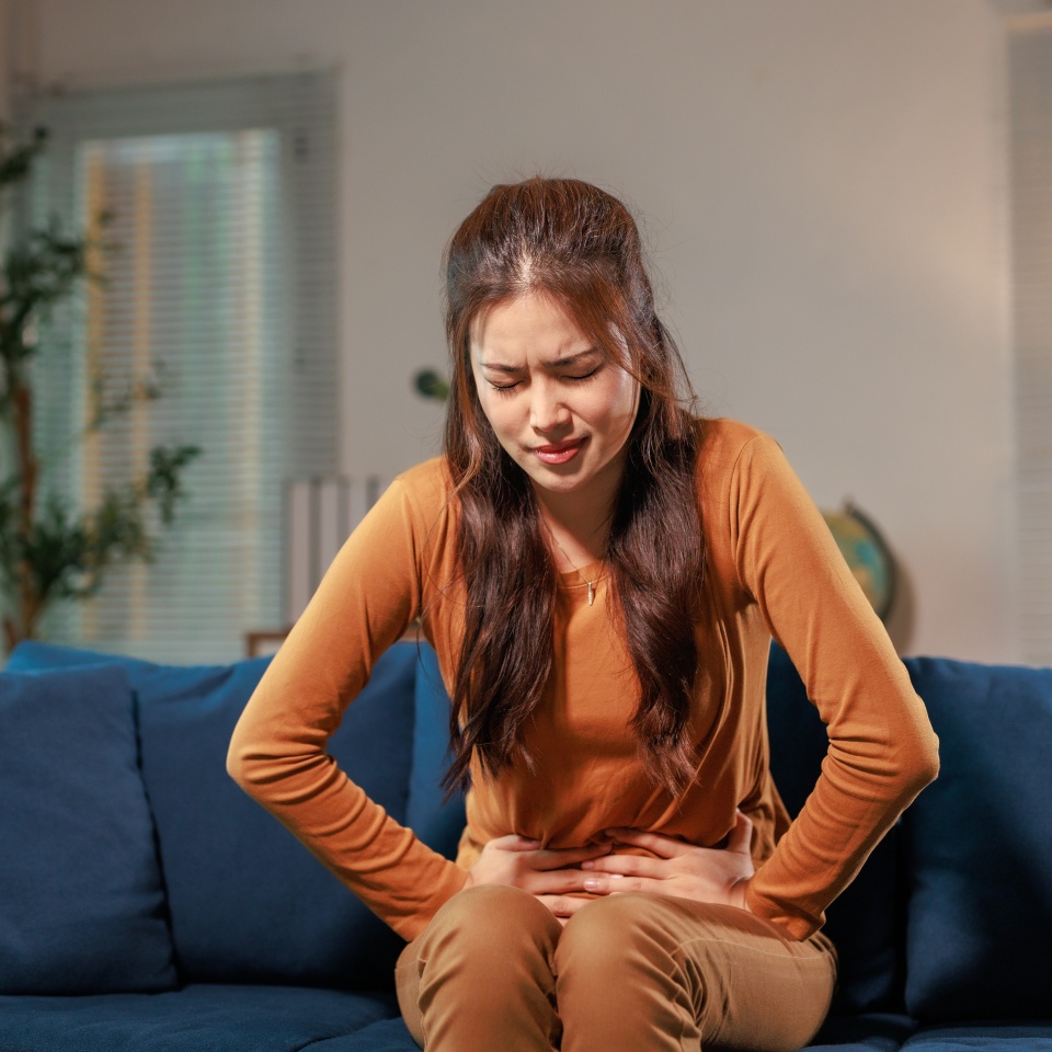 Asian woman sitting on a sofa at home, holding her belly in discomfort from severe stomach ache, experiencing symptoms of abdominal pain and cramps