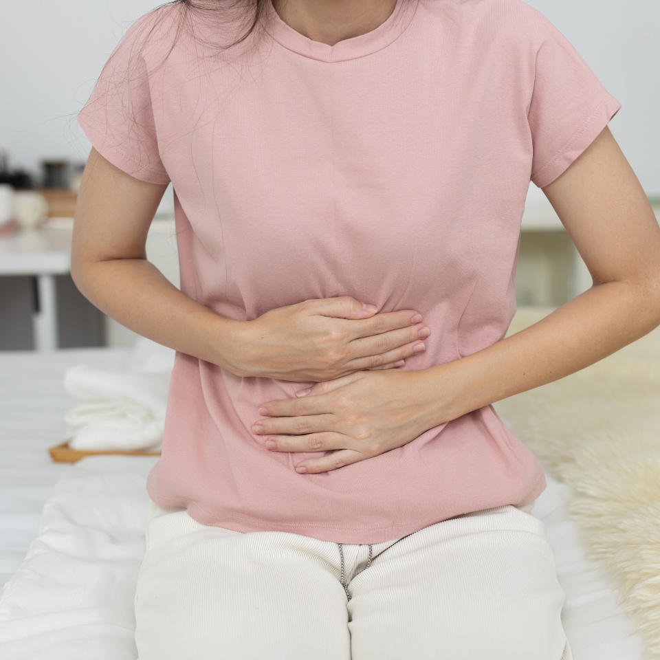 Women suffering from stomach pain or menstrual cramps while sitting on bed at home. Concept of abdominal discomfort, digestion problems or period pain in daily life.