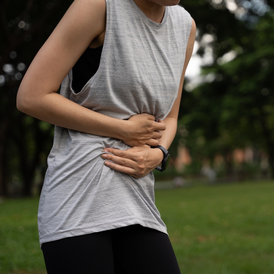 Young woman clutching stomach outdoors in pain, possibly suffering from abdominal cramps or digestive issues during exercise. Concept of health problems, stomach ache, fitness discomfort.