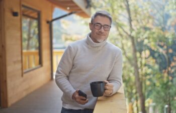 Relaxed man drinking coffee using mobile phone on balcony of modern wooden house in mountains. Portrait of happy mature businessman enjoying nature and peaceful moment of vacation, smiling.